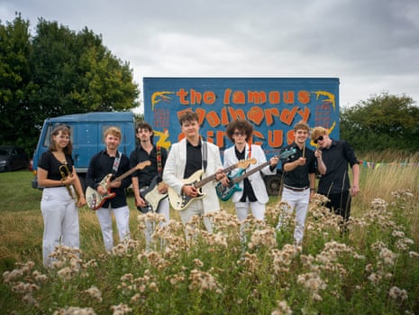 Young band members wearing white trousers and black shirts stand in a field in front of a circus van holding instruments such as guitars and a trumpet