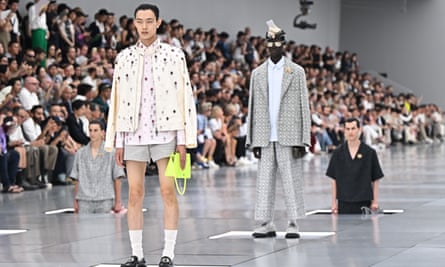 Model on catwalk wearing a t-shirt and blazer covered in gems, with shorts and high socks. Model behind wears a tweed-style suit, while two other models rise up through trapdoors behind them