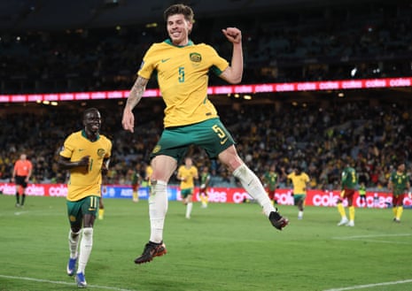 Jordan Bos of Australia jumps into the air, wearing yellow and green kit, celebrating scoring against Cameroon.