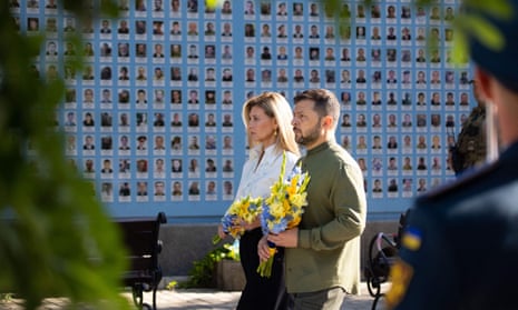 Volodymyr Zelenskiy and Olena Zelenska carry bouquets of yellow and blue flowers next to a wall covered with photographs of people