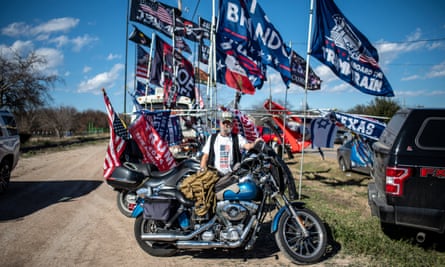 A man with a white T-shirt reading ‘God’s children are not for sale’ stands behind a motorcycle as several pro-Trump flags surround him