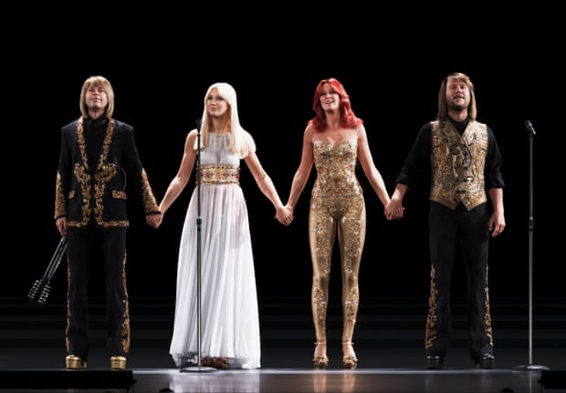 Four Abba members on stage hold hands in gold, black and white costumes