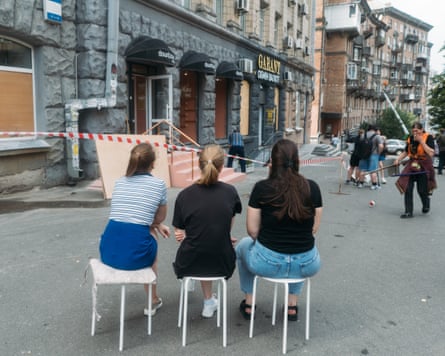 Three women sit on stools next to a cordoned-off building