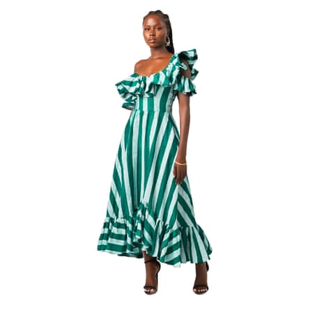 A model wears a long green and white vertical striped dress. The dress has an asymmetrical flare at the bottom and a large ruffle frill neckline that hangs off the model’s left shoulder.