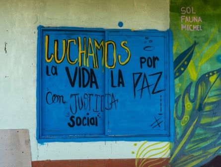 The two frames of a sliding, wooden, closed window painted bright bright, on a light green wall alongside a darker green image of a plant leaf, with the words in yellow and black: “Luchamos por la vida, la paz, con jusitica social” (We fight for life and peace with social justice).