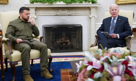 Two white mean, one younger in olive fatigues, the other elderly in a dark suit, sit in cream-colored armchairs in front of a fireplace.
