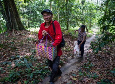 A man in a red top carries a small dog in a colourful bag while a woman following behind walks a white dog along a path