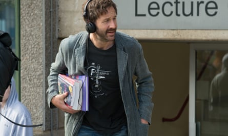 Chris O’Dowd outside a lecture theatre with headphones on in a black t-shirt and denim jacket, books under his arm, in Juliet, Naked.