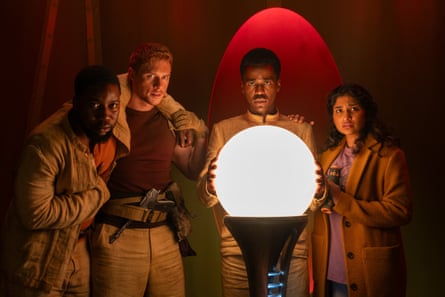 A group of four people including the Doctor and Belinda gather round a glowing orb.