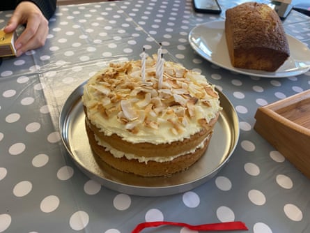 An iced cake with coconut flakes on top sitting on a tin lid with a smaller loaf cake further away on a grey tablecloth with white spots