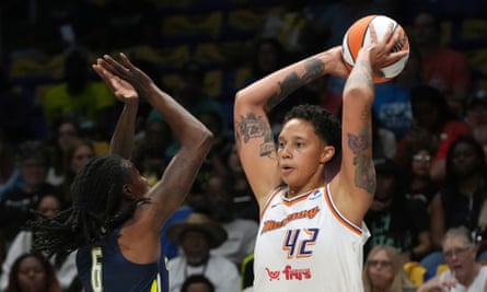 Brittney Griner: she is holding basketball over her head with both hands as if she is about to throw it; she wears a white vest with the number 44 and has heavily-tattooed arms