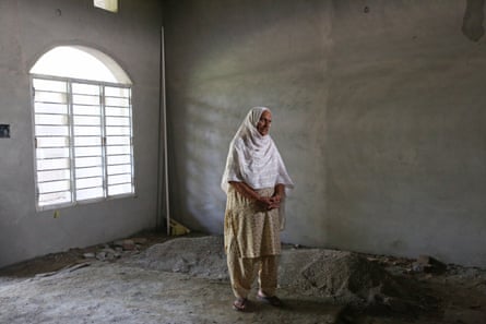 An old Indian woman in a headscarf and shalwar kameez stands in prayer in a bare room