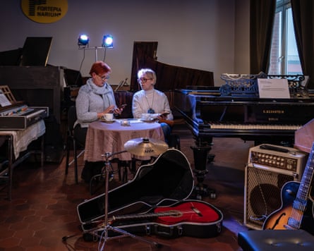 Two people sit at a small round table in a music-filled room, talking over cups of coffee, surrounded by instruments including a grand piano, guitars, and amplifiers.