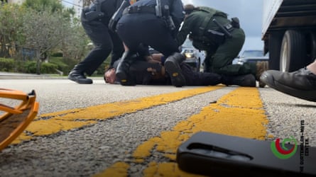 Seen from the street, a man is pinned to the ground with his hands behind his back by three black-uniformed officers