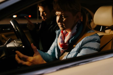 امرأة شقراء ترتدي سترة زرقاء تحمل عجلة القيادة، ورجل في مقعد الراكب