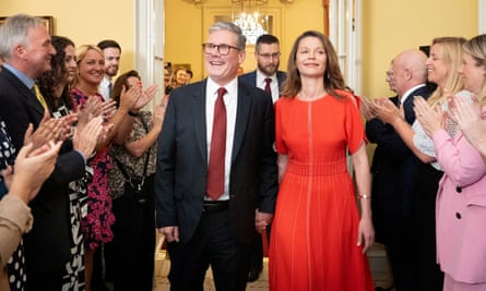 The pair flanked by clapping people. Victoria Starmer is wearing a long red dress with short sleeves