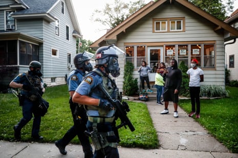 Protesters and residents watch as police in riot gear walk in front of houses