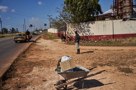 A wheelbarrow carries a bag and a sign that reads ‘carbon 1300’ by the side of the road. A man stands a few metres behind it