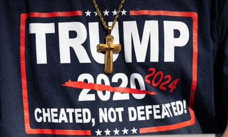 Closeup of a Trump supporter wearing a T-shirt reading Trump 2024: cheated, not defeated and a gold crucifix