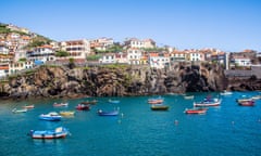 View of boats in the harbour at Câmara de Lobos on Madeira. Houses dot the coastline on a blue-sky day.