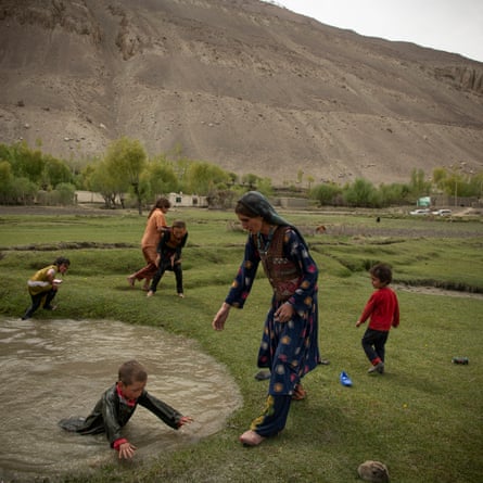 A woman reaches a hand out to a child climbing out of a pool of water. Other children play around them.