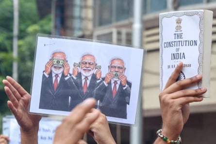 Someone holds up a copy of the constitution of India next to a poster that shows Modi covering his eyes and ears with dollar bills