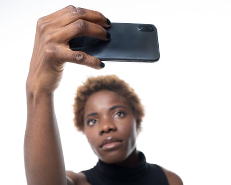 A woman with short curly hair holds up a smartphone, taking a selfie against a white background.