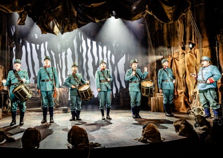 A ragtag line of soldiers are on stage, half with drums strapped over their shoulders
