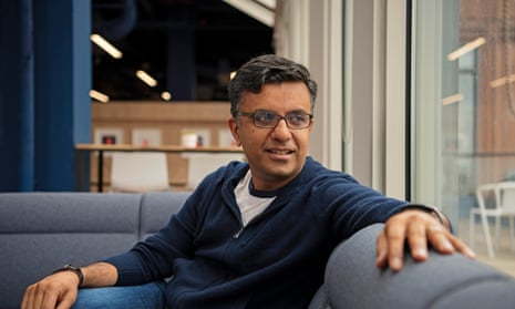 Portrait of Anil in his office, in a zip-up top and jeans, sitting on a sofa and looking over his shoulder out of the window