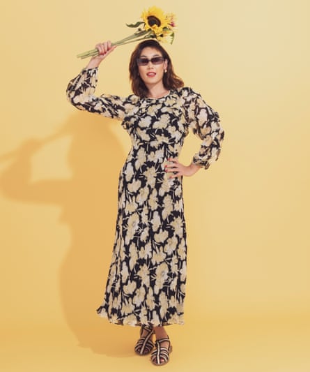 A woman stands in a black and beige floral maxidress with long sleeves. She wears black and cream sandals and holds a bunch of sunflowers above her head, and is standing in front of a yellow background