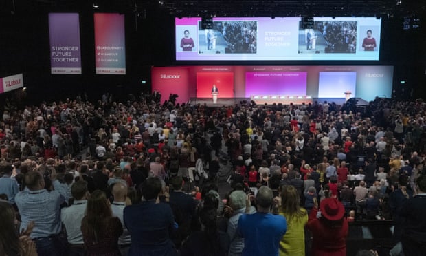 A crowd in a hall watches Keir Starmer deliver a speech in front of screensat the 2021 Labour party conference