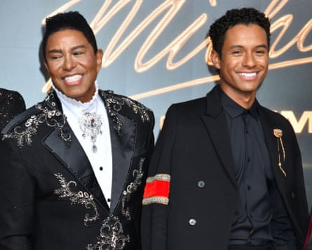 Two men in fashionable suits smiling in front of an apparently wall-size movie poster for Michael.