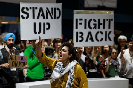 a woman wearing a keffiyeh gestures in front of people holding signs that say Stand Up, Fight Back
