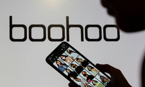 A woman poses with a smartphone showing the Boohoo app in front of the Boohoo logo.