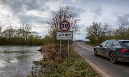 Sign for Welney below a 30mph speed-limit road sign with a river on one side and a car on the other