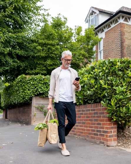 A man walking down a residential street with two canvas grocery bags in one hand and his phone in the other