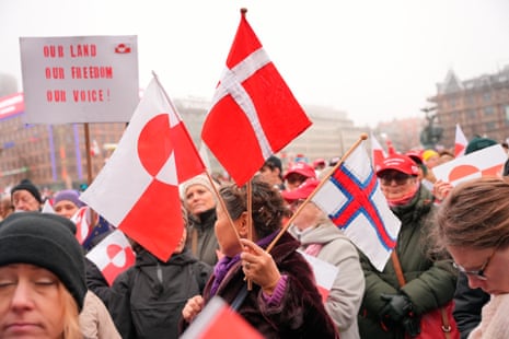 A woman holds the Greenlandic flag and the Danish flag in a crowd of protesters in Copenhagen. Behind her is a white sign with red letters reading "our land, our freedom, our voice".