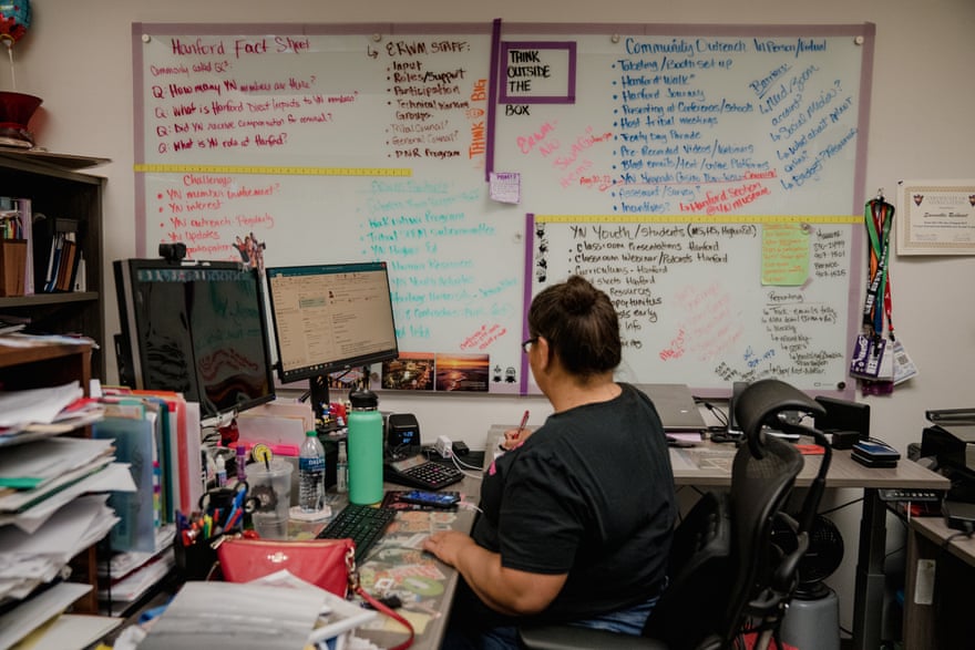 A woman is seated at a desk looking at her computer monitor. In the background, a whiteboard is filled with bullet points and lists