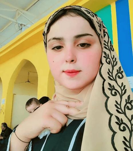 Kawthar Bashar al-Husayjawi in a patterned headscarf looks at the camera in a room with yellow walls