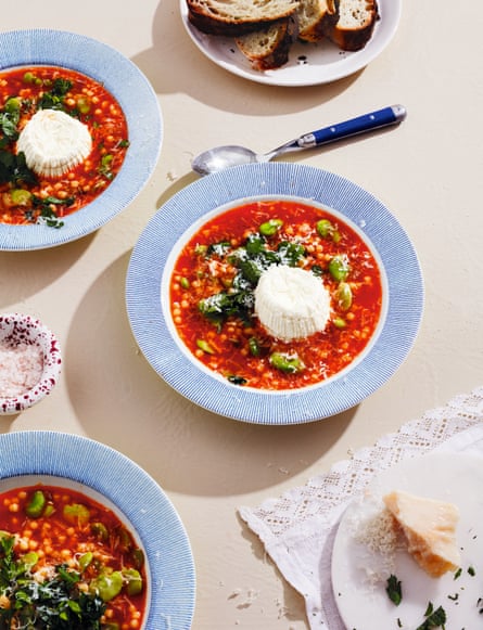 A bowl of red-coloured vegetable soup with broad beans, herbs and a ball of soft cheese in the middle
