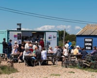 People sit at picnic benches outside a turquoise shipping container cafe and a blue shed with a 'Fresh Local Fish' sign.