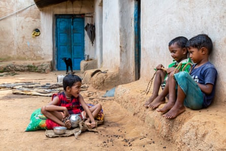 A girl sits on a cushion on the ground next to two small boys sitting on a low wall