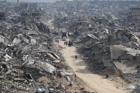 Palestinians walk through a street past destroyed buildings.