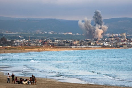 People seen on the beach in the foreground with smoke rising from an explosion in a city in the distance.
