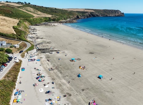 An aerial view of a beach in Cornwall, with blue water and people laid out under umbrellas on the sand.