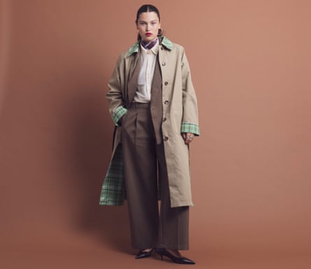 A model with slicked-back hair stands wearing a brown suit and beige trench coat, with cream shirt and high-neck patterned top underneath, plus pointy heels, rings and large hoop earrings