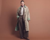 A model with slicked-back hair stands wearing a brown suit and beige trench coat, with cream shirt and high-neck patterned top underneath, plus pointy heels, rings and large hoop earrings