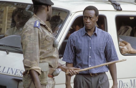 the actor Don Cheadle stands in front a minibus by a man in army fatigues with a stick