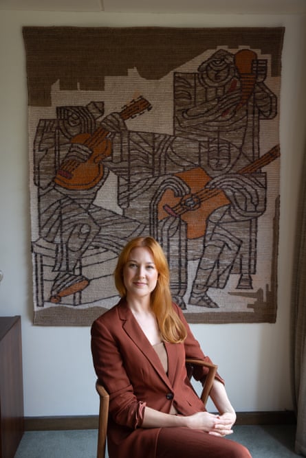A woman sitting on a chair, with a wool-weaving depicting three musicians hanging on the wall behind her.