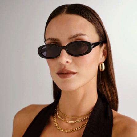 A model wears a pair of Outta Love black oval sunglasses with dark lipsick and gold jewellery.